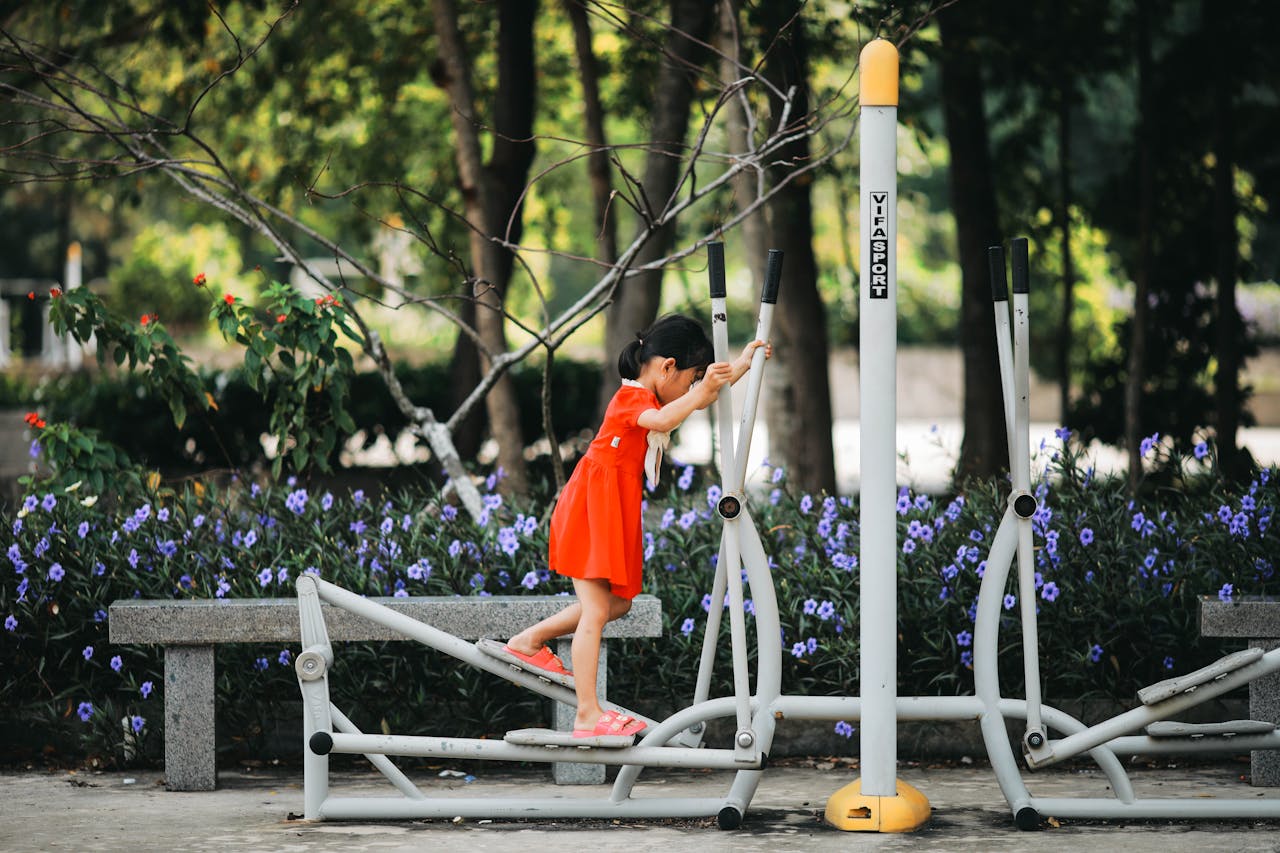services-01 A young girl in a vibrant dress uses an elliptical trainer in a lush park setting.