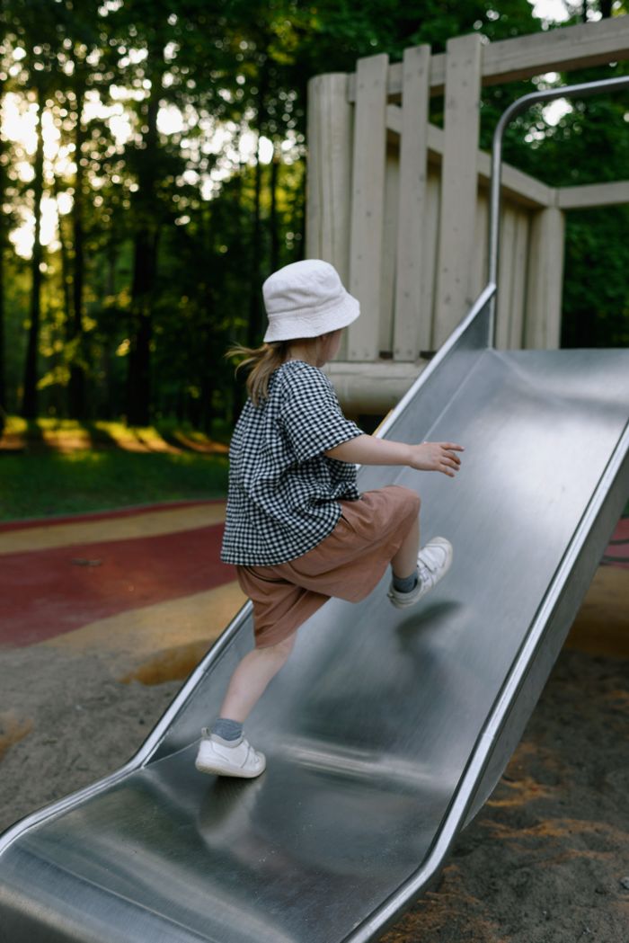 contact-img A young child with a bucket hat climbs up a slide in a sunlit outdoor playground.