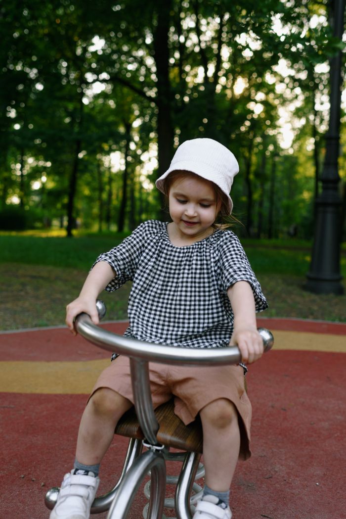 about-me-img A young girl in a hat riding a seesaw in a lush summer park.